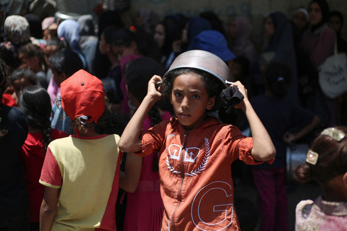 Uma menina palestina cobre a cabeça com uma panela vazia enquanto espera em um ponto de distribuição de refeições quentes em Nuseirat, no centro da Faixa de Gaza