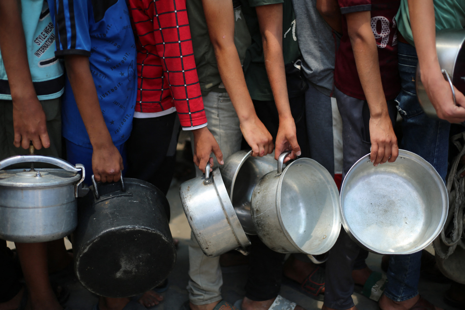 Meninos palestinos carregam panelas enquanto fazem fila em um ponto de distribuição de refeições quentes em Nuseirat, no centro da Faixa de Gaza