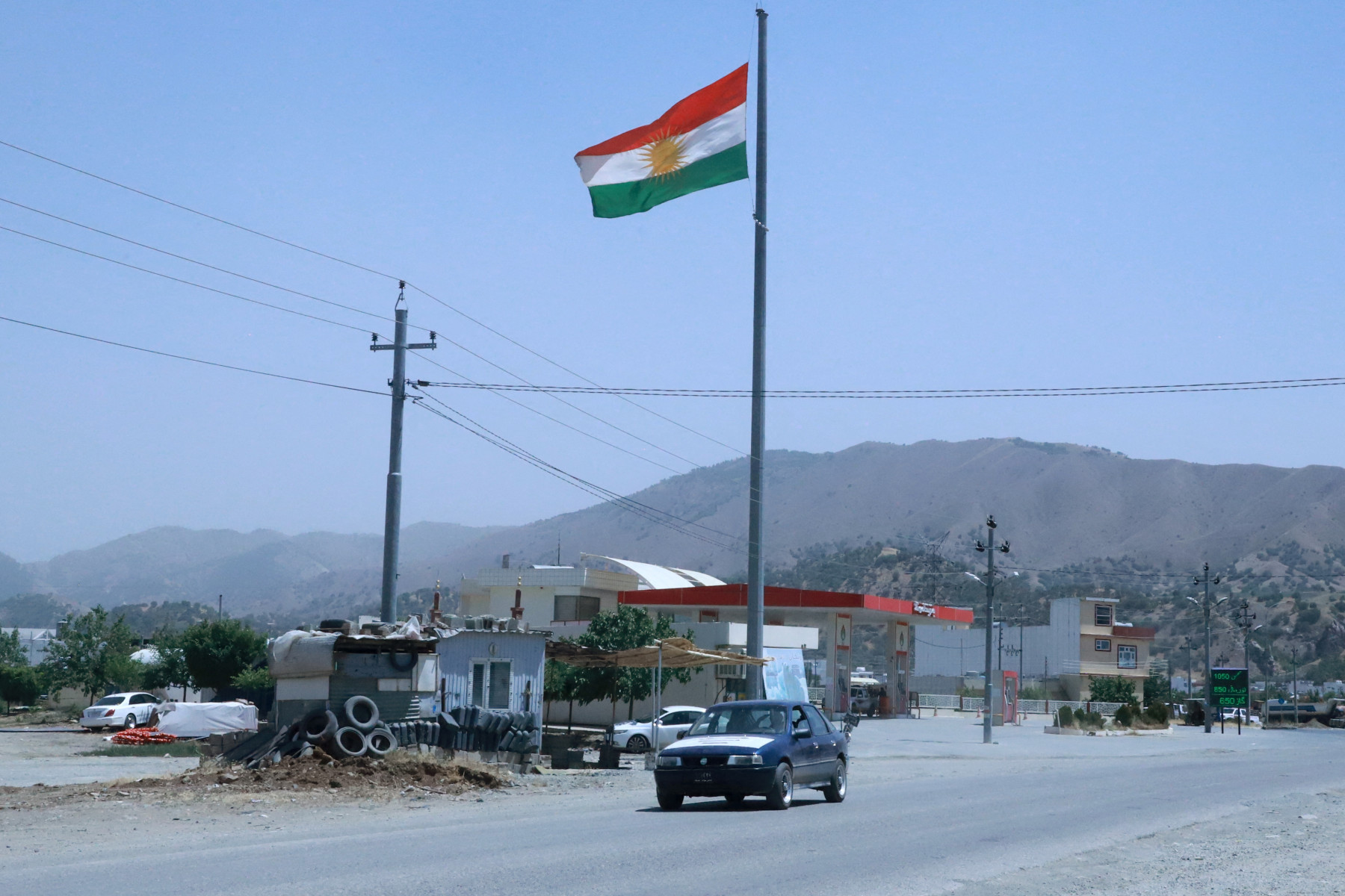 Um carro passa por um poste com uma bandeira curda na área próxima à fronteira de Bashmakh com o Irã, no distrito de Penjwin, ao norte de Sulaymaniyah, na região autônoma do Curdistão, no norte do Iraque, em 17 de junho de 2025. Perto da antes movimentada fronteira, dezenas de caminhoneiros aguardam impacientemente para retornar ao Irã, vindos da região do Curdistão iraquiano, no norte do país, trazendo não apenas suas cargas, mas, principalmente, bens essenciais para suas famílias, dias após um ataque israelense surpresa ao seu país. (Foto de Shwan MOHAMMED / AFP)
