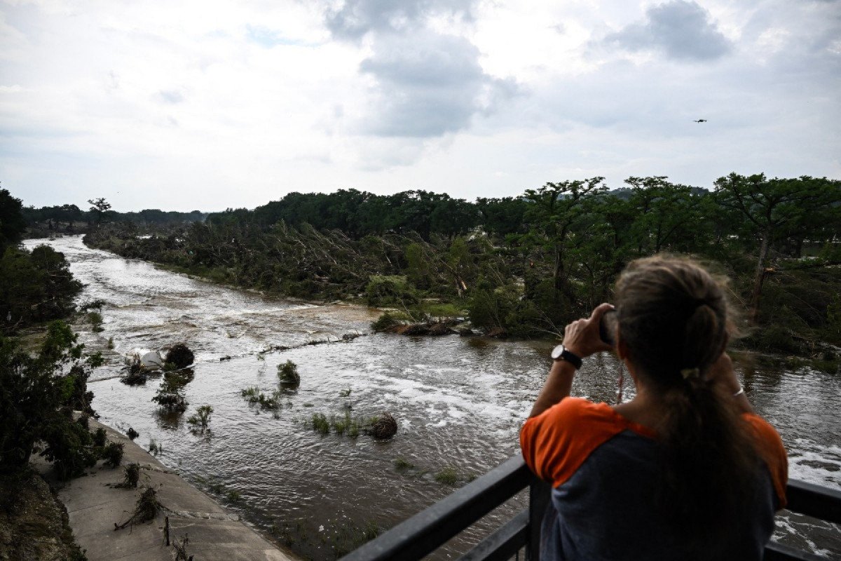 Uma mulher tira fotos do Rio Guadalupe em Kerrville, Texas, em 6 de julho de 2025, após uma grave enchente repentina que ocorreu durante o feriado de 4 de julho. Equipes de resgate no Texas correram contra o tempo no domingo para encontrar dezenas de pessoas desaparecidas, incluindo crianças, arrastadas pelas enchentes repentinas que mataram pelo menos 78 pessoas, enquanto meteorologistas alertavam para novos dilúvios. Texanos locais uniram forças com autoridades de desastres para procurar durante a noite pelos desaparecidos, incluindo 27 meninas de um acampamento de verão cristão à beira do rio. O governador do Texas, Greg Abbott, disse que o Acampamento Mystic, às margens do Rio Guadalupe, onde cerca de 750 meninas estavam hospedadas quando as águas atingiram o local, foi 