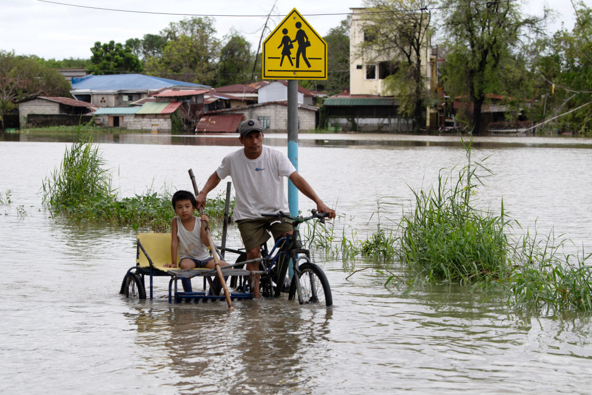 Pai e filho andam de triciclo por uma rua alagada em um vilarejo na cidade de Calumpit, província de Bulacan, ao norte de Manila, em 25 de julho de 2025, após o transbordamento de um rio devido às fortes chuvas causadas pelo Tufão Co-May. Equipes de resgate no norte das Filipinas resgataram moradores presos nas enchentes e entregaram suprimentos de barco em 25 de julho, quando o Tufão Co-May foi rebaixado para tempestade tropical horas após atingir a costa oeste. (Foto de Ted ALJIBE / AFP)