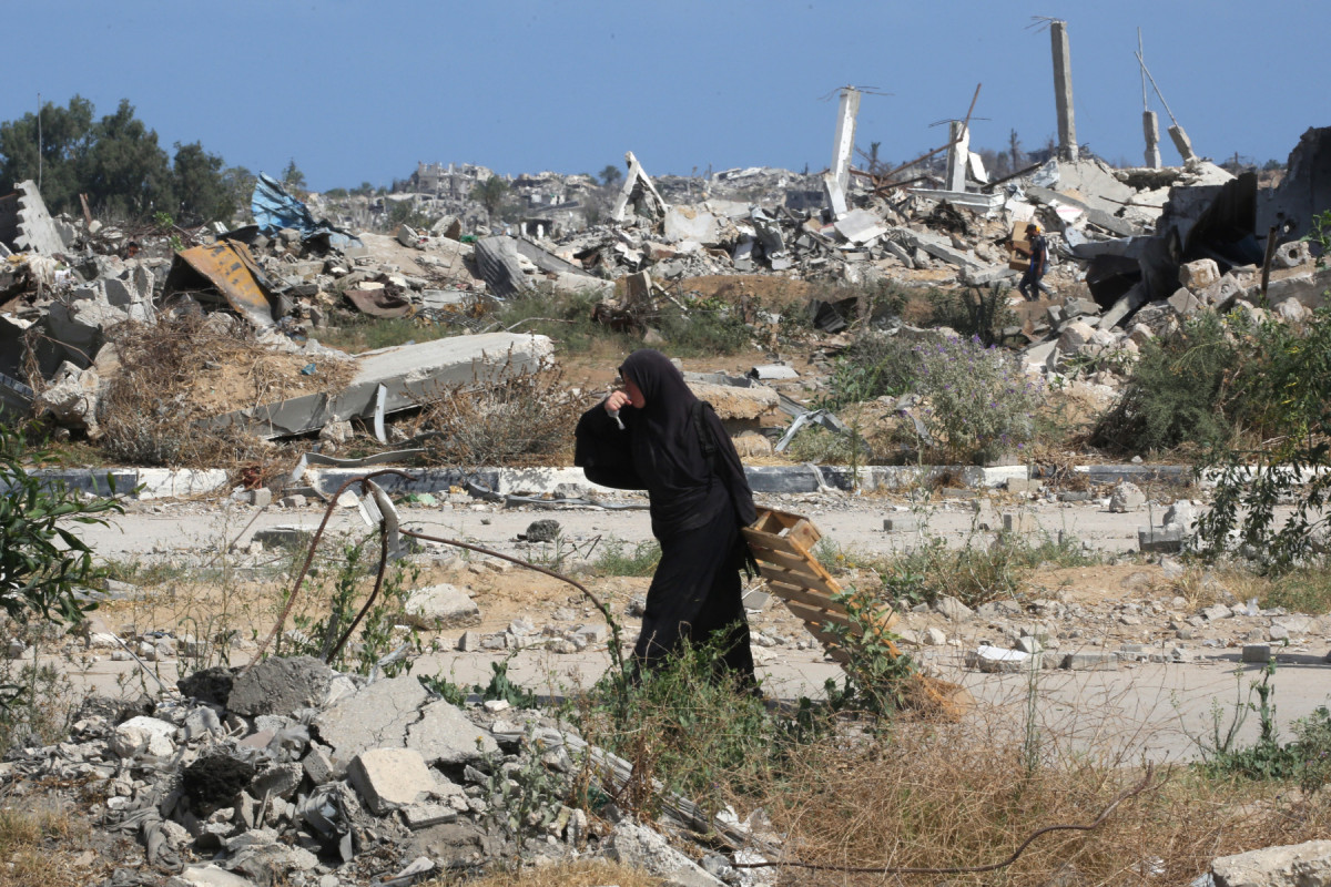 Uma mulher palestina deslocada bebe água enquanto transporta um pedaço de madeira no centro da Faixa de Gaza, em 30 de julho de 2025. Alertas de fome na Faixa de Gaza aumentaram após quase 22 meses de guerra, com o número de mortos aumentando e sem sinais de um avanço iminente nas negociações de trégua. (Foto de Eyad BABA / AFP)/ AFP