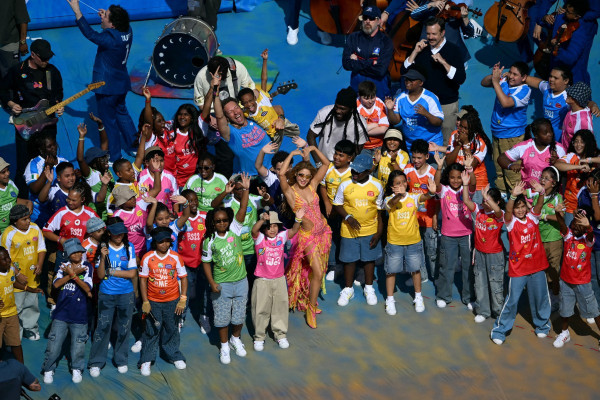A cantora, compositora e produtora colombiana Shakira (ao centro), o cantor, compositor e produtor musical nigeriano Burna Boy e o ator americano Jason Sudeikis gesticulam junto a artistas durante o show do intervalo da final da Copa do Mundo de 2026