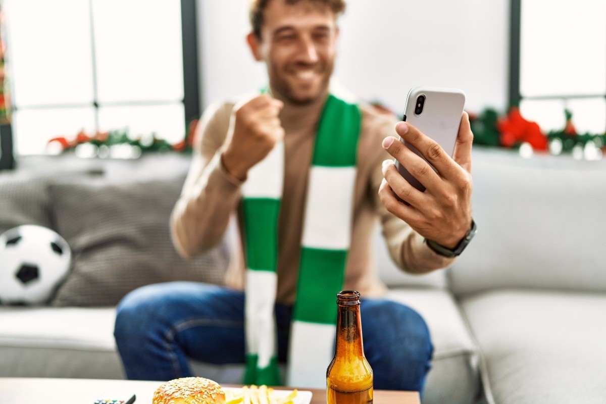 Imagem mostra homem segurando celular enquanto acompanha partida de futebol/Magnific
