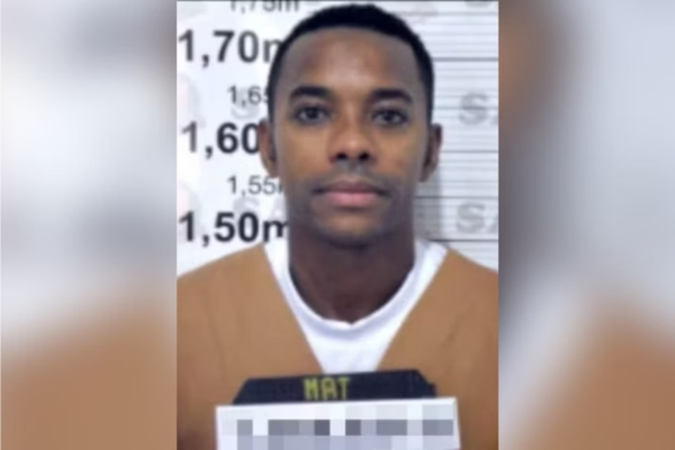 
Ex-jogador Robinho, um homem negro de cabelos curtos, vestindo uniforme de presidirio, posando para foto na cadeia (Crdito: Reproduo)